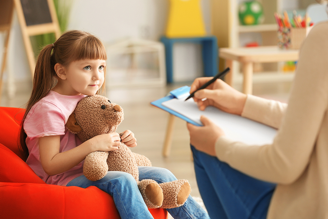 Young girl around five years holding teddy bear on lounge looking at counsellor who is scribing